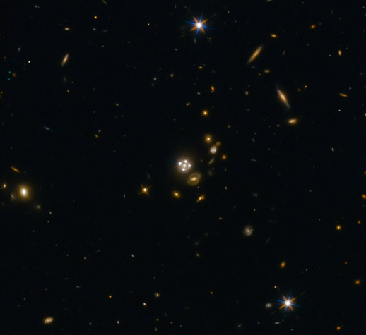 Rysunek 1. W centrum zdjęcia widzimy Kwazar poddany soczewkowaniu grawitacyjnemu przez galaktykę znajdującą się przed nim. Cztery równo ułożone punkty są obrazami tego samego Kwazara, powstałe poprzez grawitacyjne zakrzywianie promieni światła przez soczewkującą galaktykę. Źródło: ESA/Hubble, NASA, Suyu et al. (Lensed quasar and its surroundings | ESA/Hubble (esahubble.org))